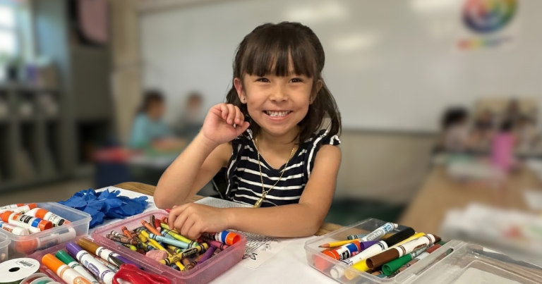 Sophia smiling at her classroom desk surrounded by art supplies and crayons
